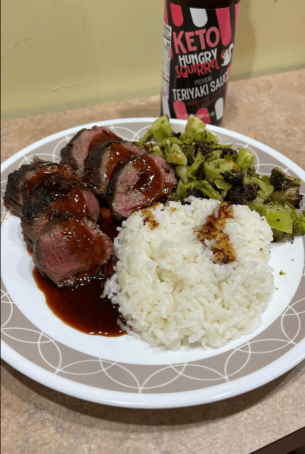 A plate of Seared Teriyaki Glazed Venison Backstrap, with rice and brocolli. - Hungry Squirrel Teriyaki Sauce on the background.- From Instagram follower:  @jimmydean1147